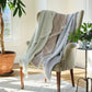 A beige armchair draped with three John Robshaw Lavanya Throw Collection cotton blankets in pastel shades sits by a potted plant and a small table with purple flowers, bathed in sunlight.