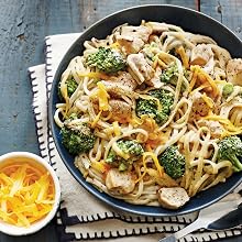 Enjoy a bowl of Super Easy Pasta! by Simon & Schuster, featuring tender pasta with grilled chicken, broccoli, and shredded cheese—served on a dark wooden table with a small bowl of extra cheese on the side. Perfect for anyone seeking easy pasta recipes or quick pasta dishes for a satisfying meal.