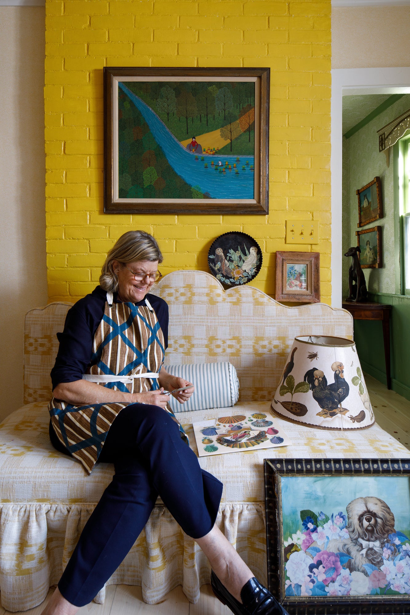 A woman sits on a patterned couch amid craft supplies, art, and decor in a bright room, evoking the creativity of "A Découpage Workshop With Sister Parish Design" by Hive Floral Studio.