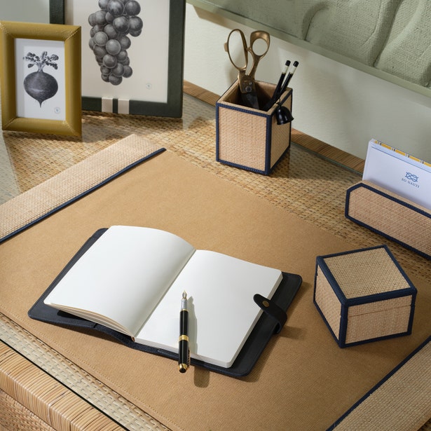 A tidy desk setup featuring the Pigeon & Poodle Hanford Desk Accessory Collection, an open notebook, pen, framed photos, pen holders, and a notepad arranged on a woven raffia and leather mat.