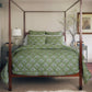 A neatly made four-poster bed features the Jeeva Sage Bedding Collection by John Robshaw, flanked by matching bedside tables, a lamp, a vase with branches, and a framed picture on the wall.