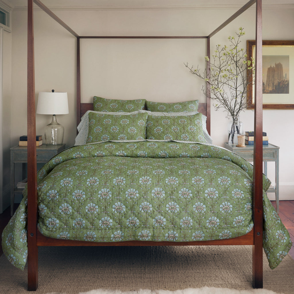 A neatly made four-poster bed features the Jeeva Sage Bedding Collection by John Robshaw, flanked by matching bedside tables, a lamp, a vase with branches, and a framed picture on the wall.