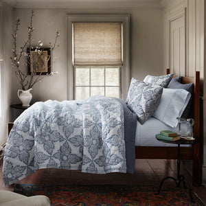 A wooden bed with blue-and-white Dasati Lapis King Shams from HIVE Home, Gift and Garden is set near a window with a woven shade; books, a glass pitcher, and a glass sit on a small side table.