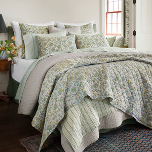 A bright, cozy bedroom with white walls features a neatly made bed adorned with the John Robshaw Tiya Periwinkle King Quilt—hand-quilted and floral-patterned—plus multiple pillows by the window.