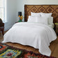 A neatly made bed featuring the John Robshaw Vivada Bedding Collection, dressed in crisp white bedding and textured pillows, stands against a woven headboard. A colorful rug and a small nightstand with flowers add charm to the cozy, well-lit bedroom. Layered with soft, reversible quilts from the Vivada collection, the space feels both inviting and effortlessly elegant.