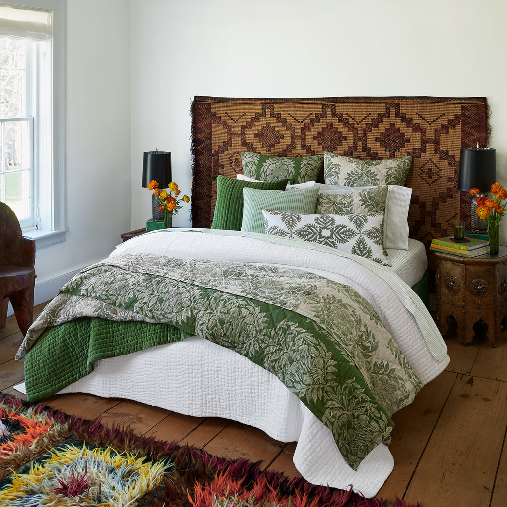 A neatly made bed with green and white botanical cotton linen blend bedding, a John Robshaw Verdin Dark Sage Bolster Pillow, decorative pillows, a patterned headboard, and side tables with black lamps and orange flowers.