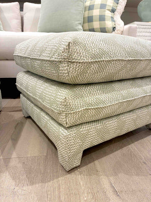 The Empire Ottoman by Bunny Williams, featuring a patterned beige upholstery and two stacked cushions, rests on wooden flooring with part of a sofa and pillows seen in the background.