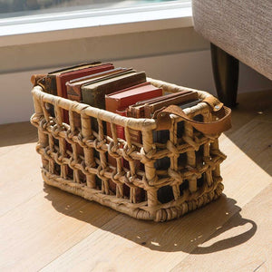The Small Somerset Basket by Pigeon & Poodle, featuring leather handles, holds several hardcover books and sits on a wooden floor near a window and sofa.