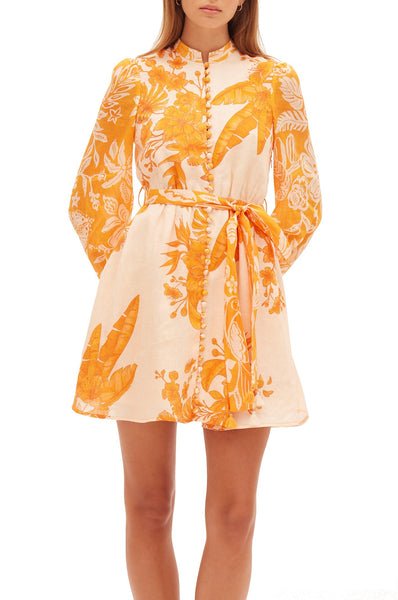 Woman wearing a long-sleeve, button-down Kate Ford Hester Mini Dress with a vibrant orange leaf and floral spliced print design. The dress features a self waist belt and ends above the knee.