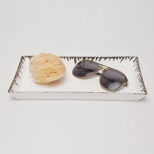 The Pigeon & Poodle Rio Silver and White Tray, a rectangular porcelain piece with metallic edges, holds a natural sea sponge and black sunglasses.
