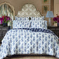 A neatly made bed features the Kanak Indigo Bedding Collection by John Robshaw, with hand-quilted blue and white floral cotton, a patterned headboard, matching side tables and lamps, plus vases of purple flowers on either side.
