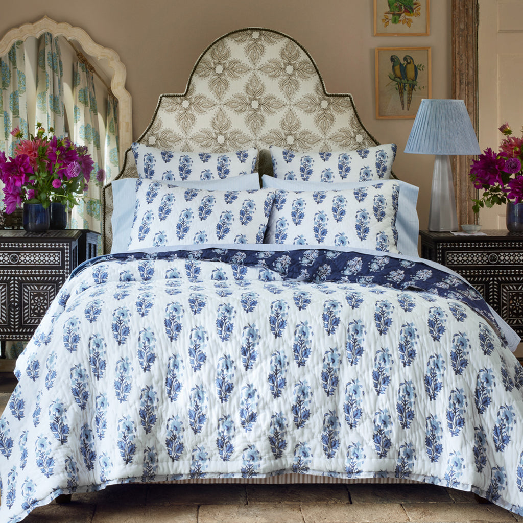 A neatly made bed features the Kanak Indigo Bedding Collection by John Robshaw, with hand-quilted blue and white floral cotton, a patterned headboard, matching side tables and lamps, plus vases of purple flowers on either side.