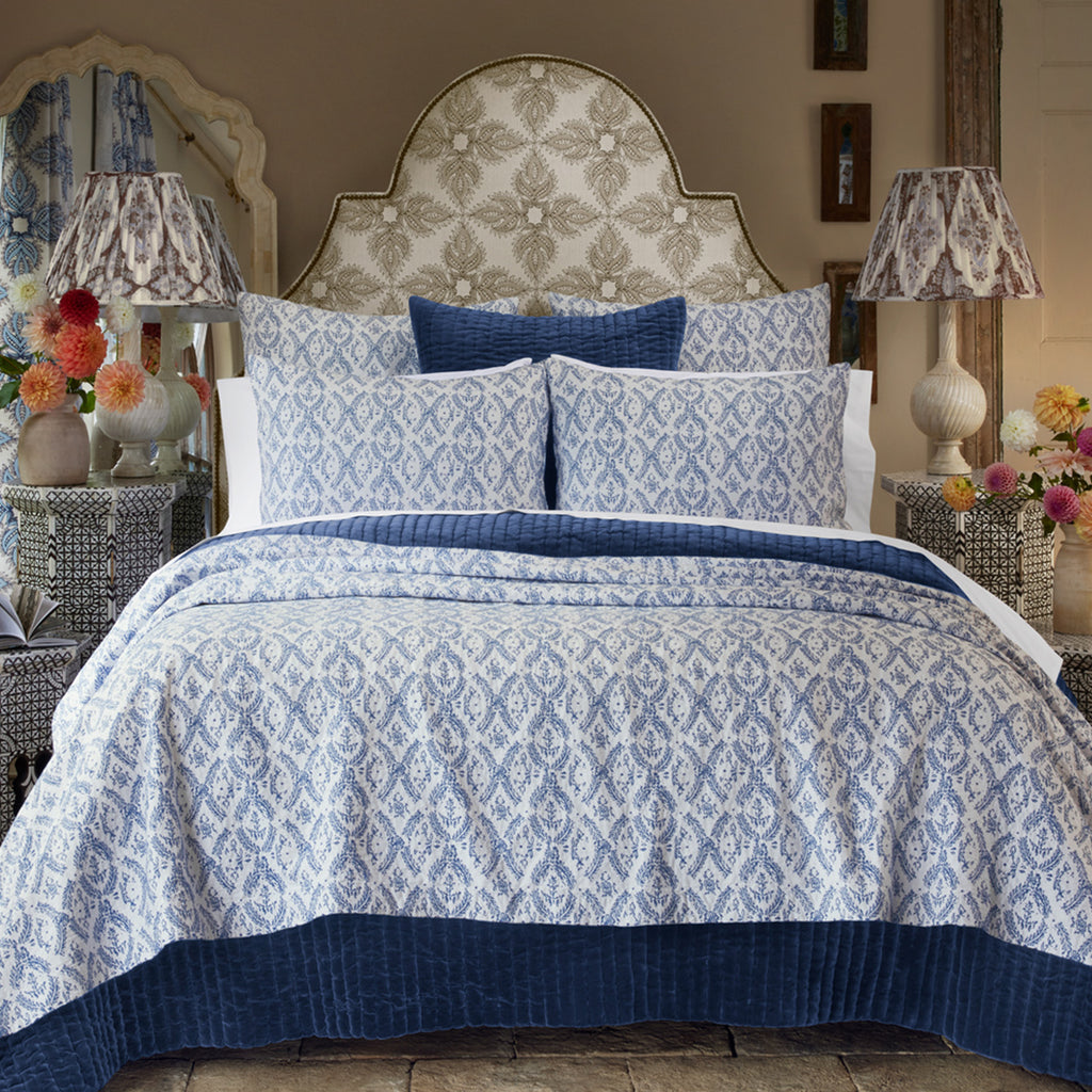 The Aashna Azure Bedding Collection by John Robshaw features blue and white cotton jacquard bedding with matching pillows, a decorative headboard, and is paired with bedside tables topped with lamps and vases of flowers.