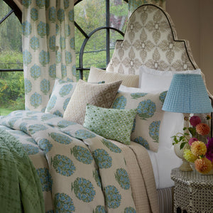 A neatly made bed adorned with the John Robshaw Vani Leaf King Duvet Set, showcasing blue and green floral motifs, complemented by matching pillows, a decorative headboard, a side table with a blue lamp, and a vase of flowers placed by the window.