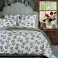 A bed with a scalloped patterned headboard features the John Robshaw Farah Light Indigo Bedding Collection, showcasing hand quilted white fabric with blue and green floral prints. A bedside table displays a vase of red flowers and a tissue box.