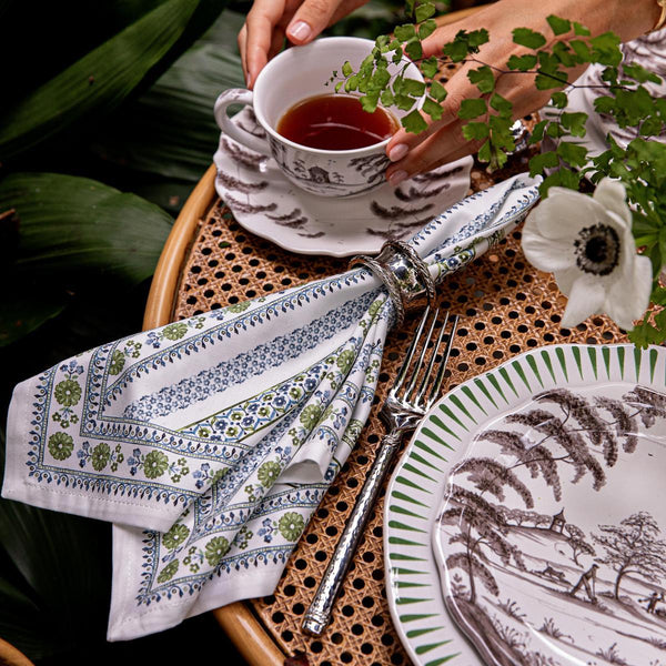 A person holds a teacup over a table featuring Juliska’s Seville Green Stripe Table Linen Collection, with patterned plates, a floral napkin in a ring, a fork, and greenery decorating the background.