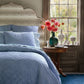 The Nandi Bedding Collection by John Robshaw, featuring light blue hand-quilted cotton quilts, dresses the bed that stands beside a round wooden table holding a vase of pink flowers, set near a large window with a garden view.