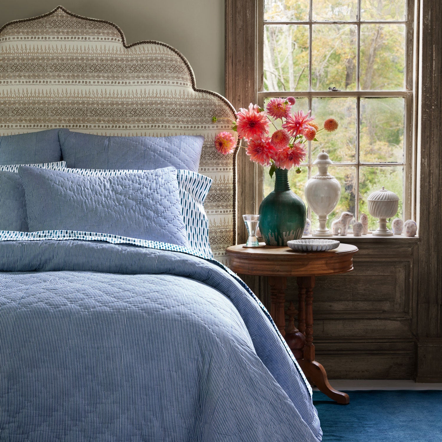 The Nandi Bedding Collection by John Robshaw, featuring light blue hand-quilted cotton quilts, dresses the bed that stands beside a round wooden table holding a vase of pink flowers, set near a large window with a garden view.