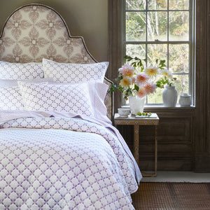 A neatly made bed with a patterned headboard features the John Robshaw Layla Lavender Coverlet; white cotton voile bedding with purple accents sits by a window, while a side table holds flowers and decor inspired by Mughal gardens.