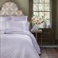A neatly made bed with the John Robshaw Nandi Lavender Coverlet and pillows stands by a window, beside a table topped with a vase of pink and white flowers.