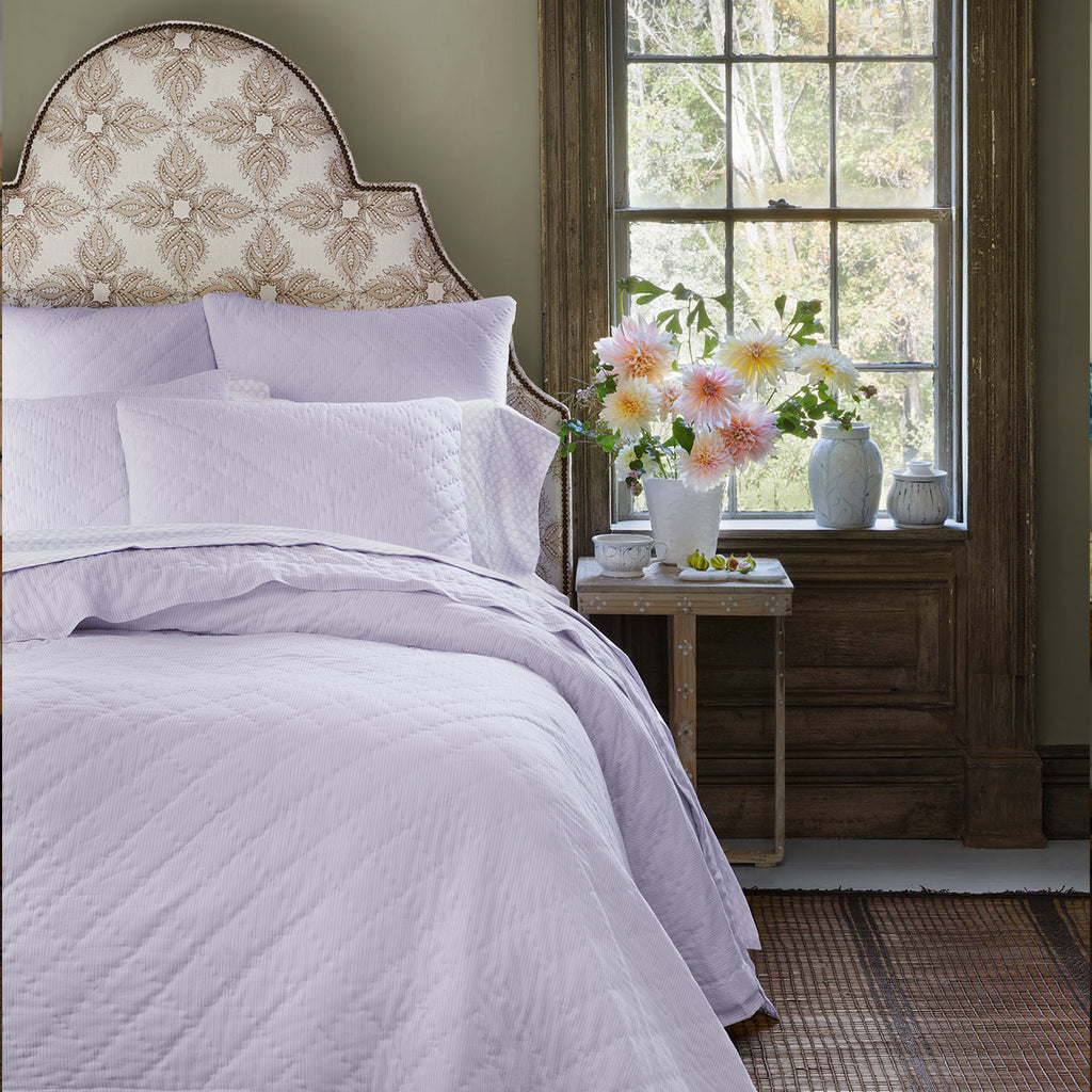 A neatly made bed with the John Robshaw Nandi Lavender Coverlet and pillows stands by a window, beside a table topped with a vase of pink and white flowers.
