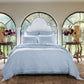 A neatly made bed with the John Robshaw Nandi Light Indigo Quilt is centered between two wooden side tables with vases of flowers, in a room featuring arched windows that overlook a garden.