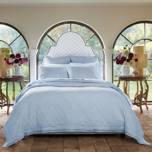 A neatly made bed with the John Robshaw Nandi Light Indigo Quilt is centered between two wooden side tables with vases of flowers, in a room featuring arched windows that overlook a garden.