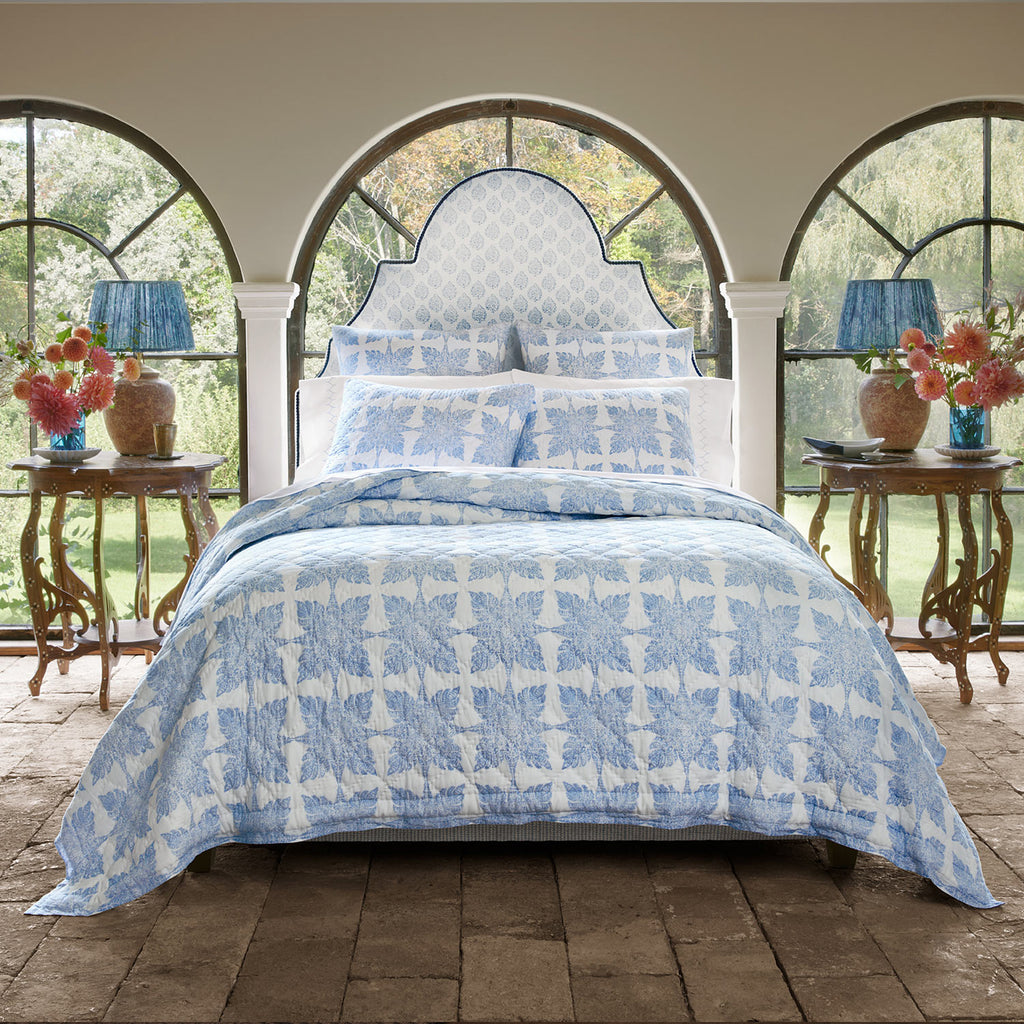 A bedroom with the John Robshaw Jaya Azure Bedding Collection—blue and white block printed cotton voile—on the bed, blue lamps and flowers on side tables, set before large arched windows.