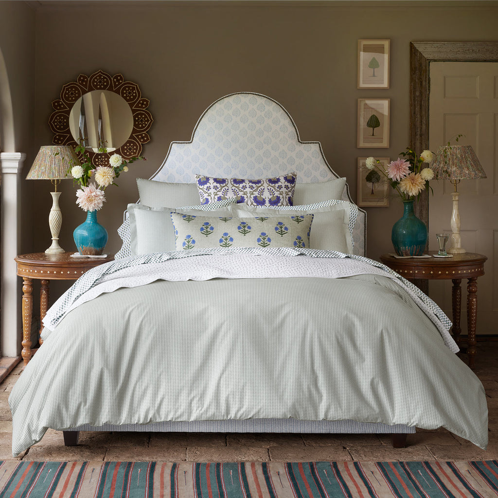A neatly made bed featuring the John Robshaw Cinde Sage Bedding Collection is flanked by wooden nightstands with blue glass lamps and flowers in a bright, traditionally decorated bedroom.