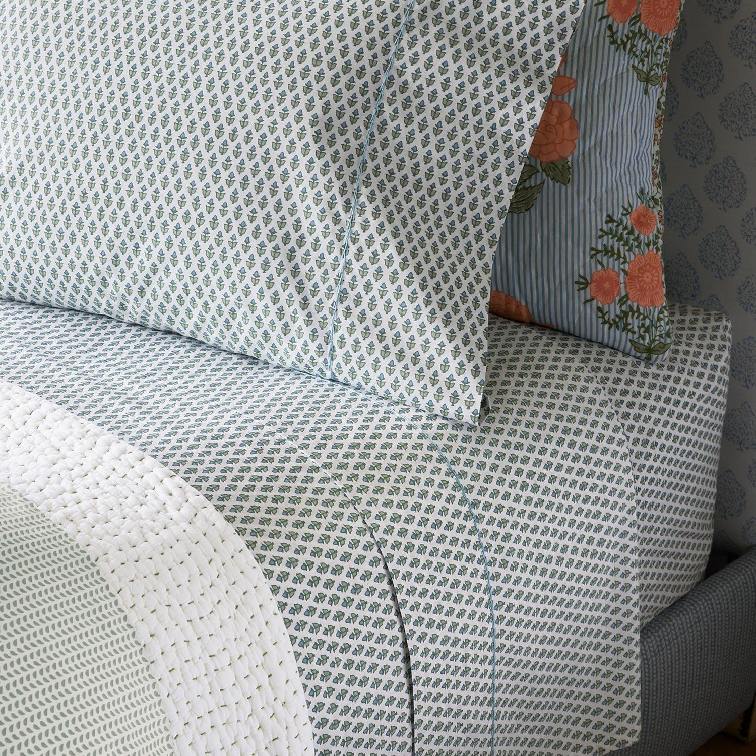 The Cala Sage Bedding Collection by John Robshaw features 400 thread count organic cotton sheets and pillowcases in a light blue and white pattern, plus a striped block print bouquet pillow and a textured white blanket at the bed's foot.