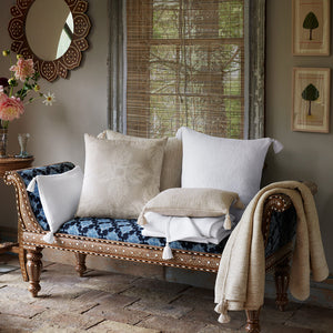 A carved wooden bench with patterned blue upholstery is topped with the Verdin Sand Pillow by John Robshaw, beige and white throws, while a mirror, flowers, and framed art—including a subtle nazar motif—decorate the cozy room.