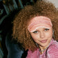 A person with curly hair sits on a dark chair, wearing the Slip Silk Sleep Mask in pink on their forehead and a pink velvet top, ready for beauty sleep.