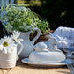 The Whitewash Bunny Serveware Collection by Juliska features a ceramic butter dish with a rabbit lid, showcased alongside elegant tableware, sparkling glassware, wildflowers in a jug, and a daisy-filled pitcher.