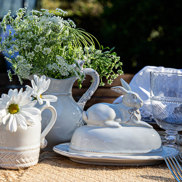 The Whitewash Bunny Serveware Collection by Juliska features a ceramic butter dish with a rabbit lid, showcased alongside elegant tableware, sparkling glassware, wildflowers in a jug, and a daisy-filled pitcher.