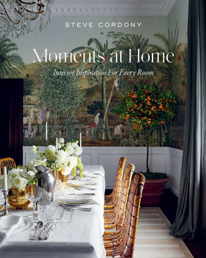 Elegant dining room styled in the signature aesthetic of Steve Cordony, featuring a long table set for a meal, rattan chairs, white flowers, and a large potted orange tree, all against a scenic mural—refined home styling and inspired interior design as showcased in “Moments At Home” by Chronicle Books.