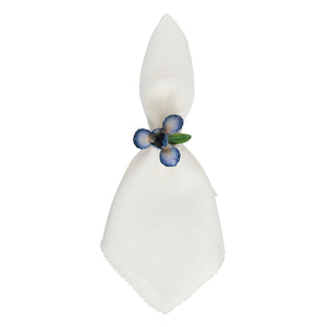 A white cloth napkin is folded vertically and held by the Juliska Meadow Walk Iris Napkin Ring Set, featuring blue and purple artificial flowers with green leaves—ideal for a spring tablescape.