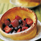 A golden Dutch baby pancake brimming with strawberries, raspberries, and blueberries, finished with a sprinkle of powdered sugar and presented on a white plate—a deliciously simple creation inspired by the Barefoot Contessa Back To Basics cookbook from Penguin Random House.