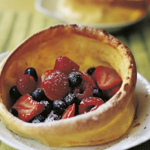 A golden Dutch baby pancake brimming with strawberries, raspberries, and blueberries, finished with a sprinkle of powdered sugar and presented on a white plate—a deliciously simple creation inspired by the Barefoot Contessa Back To Basics cookbook from Penguin Random House.
