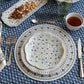 A dining table set with patterned plates, cutlery, a glass, tea, and a nut platter is arranged on a blue and white floral tablecloth, accented by the cork-backed Seville Placemat from Juliska.