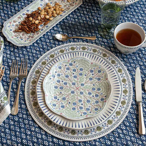 A dining table set with patterned plates, cutlery, a glass, tea, and a nut platter is arranged on a blue and white floral tablecloth, accented by the cork-backed Seville Placemat from Juliska.