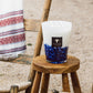 Large Baobab Collection Feathers Touareg scented candle with three wicks on a wooden stool outdoors, next to a rustic chair with a patterned white throw.