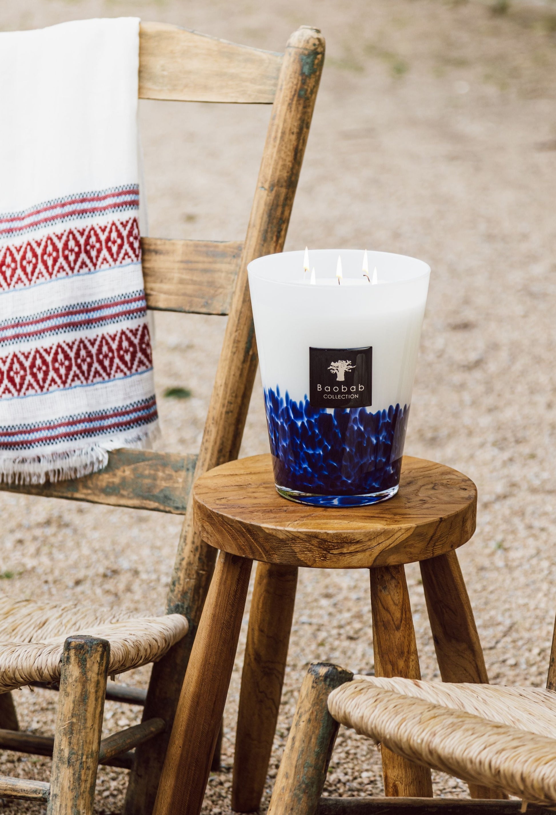 Large Baobab Collection Feathers Touareg scented candle with three wicks on a wooden stool outdoors, next to a rustic chair with a patterned white throw.