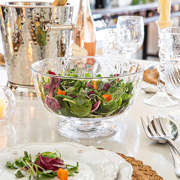 A clear mouth-blown glass bowl from Juliska’s Puro Glass Serveware Collection holds mixed greens and orange veggies, its textured lines catching the light on a set dining table with glasses, plates, and an ice bucket behind.