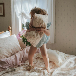 A young child stands on a bed, playfully holding the Gooselings Bows & Butterfly Kisses Toddler Pillow in front of their face, surrounded by softly lit patterned bedding and pale wallpaper—ready for sweet dreams.