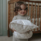 A young child stands in front of a wooden crib, holding two Gooselings Animal Parade Ivory Toddler Pillows and smiling—ready for sweet dreams.
