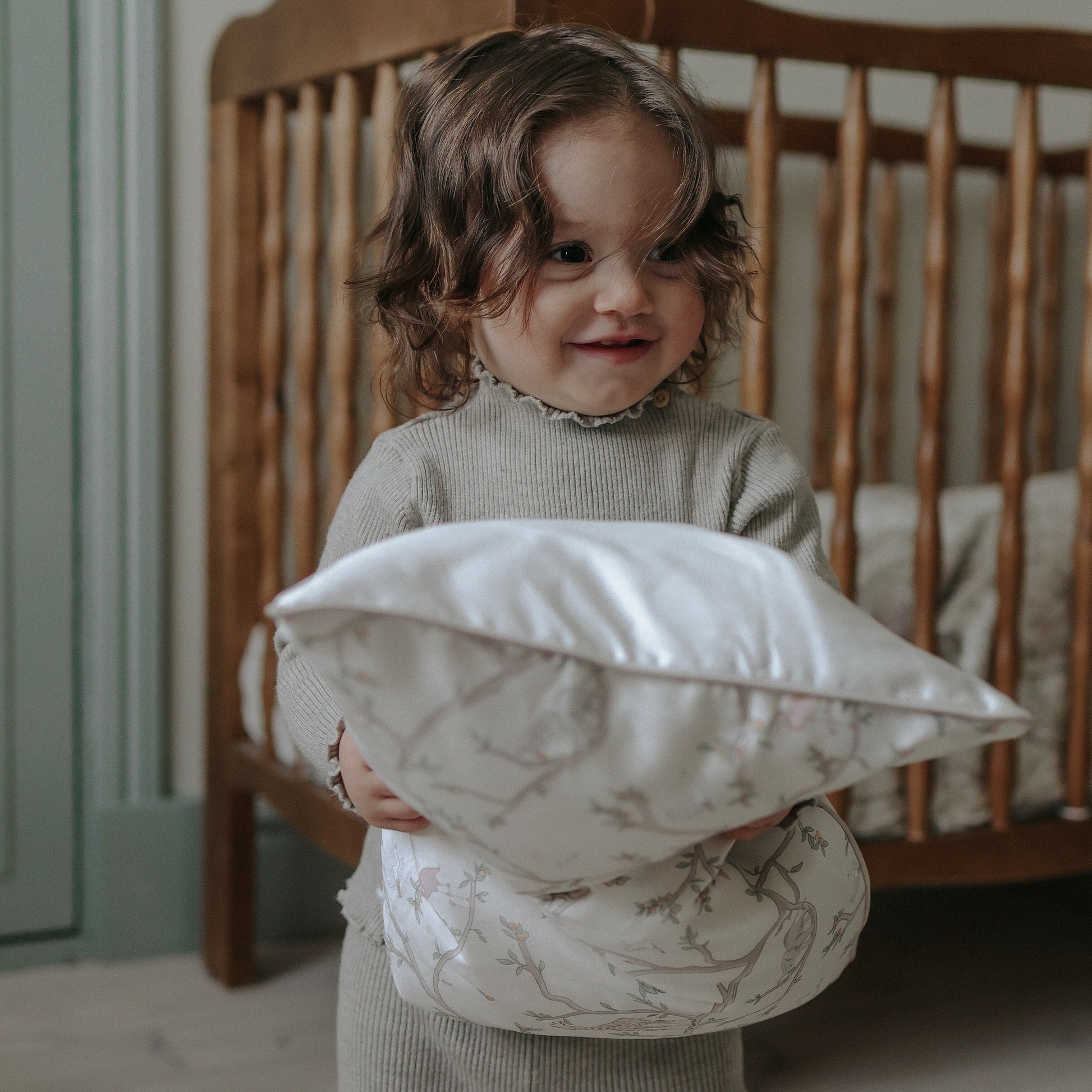 A young child stands in front of a wooden crib, holding two Gooselings Animal Parade Ivory Toddler Pillows and smiling—ready for sweet dreams.