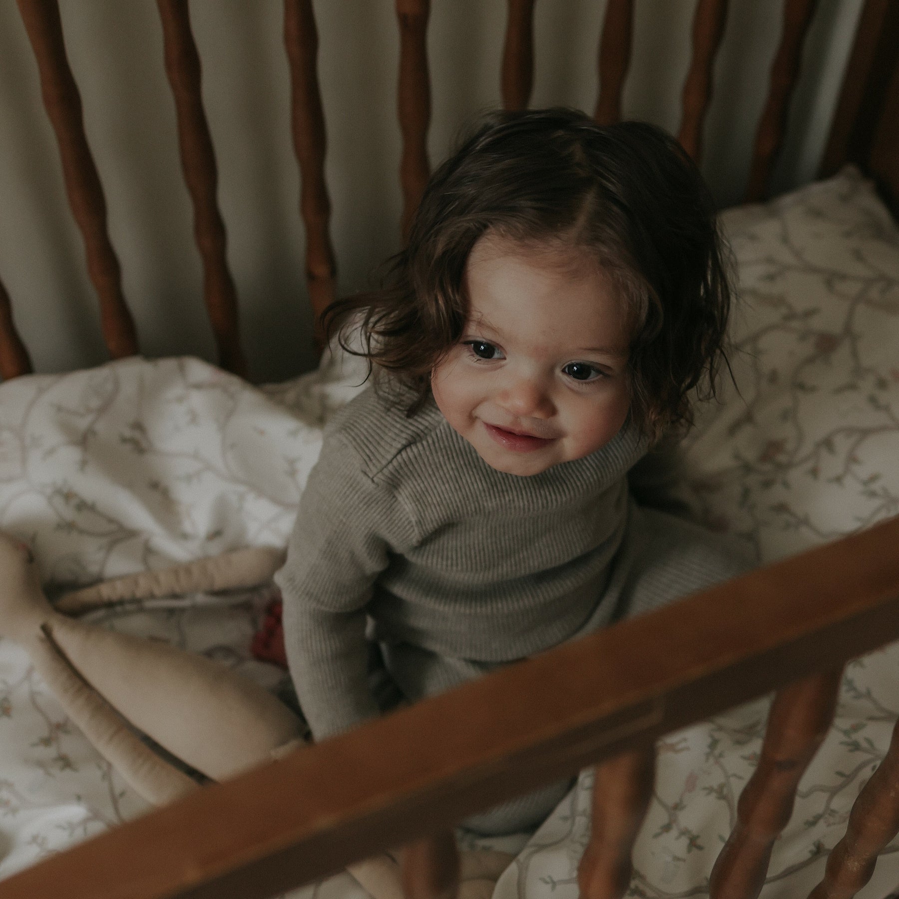 A young child with wavy brown hair sits in a crib, smiling in a grey outfit. Beside them is a soft doll on the Gooselings Animal Parade Ivory Crib Sheet, crafted from extra soft OEKO-TEX certified cotton sateen with patterned bedding.