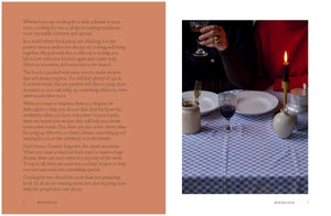 A woman in a dark sweater sits at a table set for “A Meal For Two” by Chronicle Books, featuring a blue checkered tablecloth, wine, cutlery, plates, and a candle, beside another person dressed in red.
