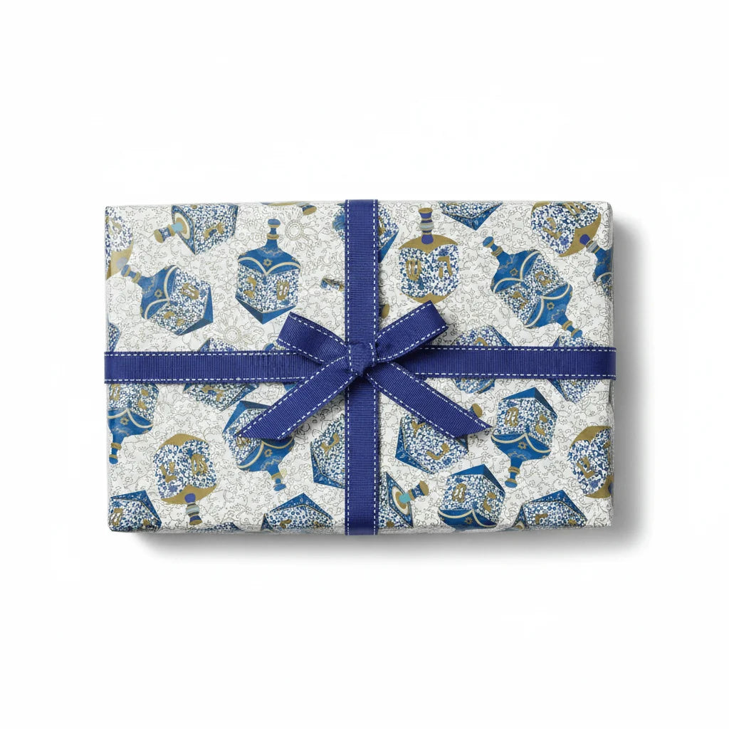 A rectangular gift from Caspari’s Patterned Dreidel Collection, wrapped in white paper adorned with a blue and gold dreidel design and finished with a blue ribbon and bow, makes the perfect Hanukkah holiday present, shown on a white background for a festive touch.