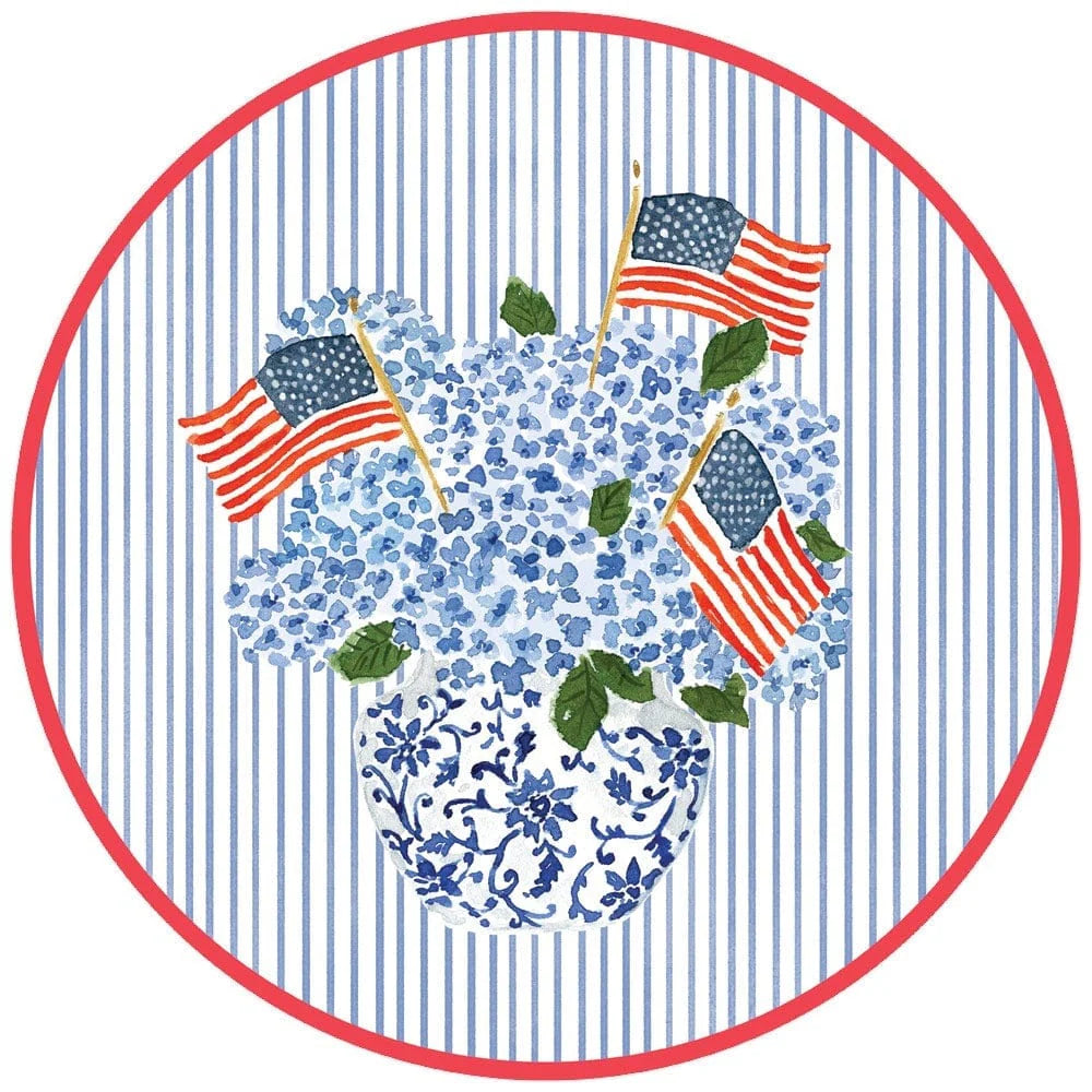 The Flags And Hydrangeas Collection by Caspari features blue hydrangeas and small American flags in a floral vase, set on a picnic table with blue stripes and a red border—ideal for summer celebrations.